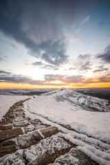 Mountain path in winter up in the Debyshire Peak District hills. Deep snow with clear stone path ahead leading the way. Beautiful orange sunrise breaking through clouds and icy weather.