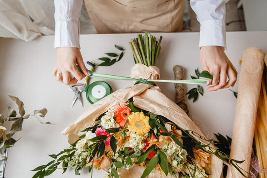 a female florist ties a green ribbon bow on a bouquet of flowers wrapped in craft paper on the desktop. Top view.