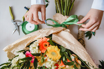 a female florist ties a green ribbon bow on a bouquet of flowers wrapped in craft paper on the desktop. Top view. - obrazy, fototapety, plakaty