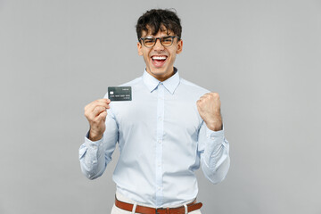Young fun rich smiling employee business latin man corporate lawyer 20s wearing classic white shirt glasses hold in hand credit bank card do winner gesture isolated on grey background studio portrait