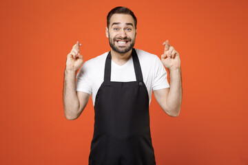 Young man 20s barista bartender barman employee wear black apron white t-shirt work in coffee shop keeping fingers crossed make wish eyes closed isolated on orange background Small business startup.
