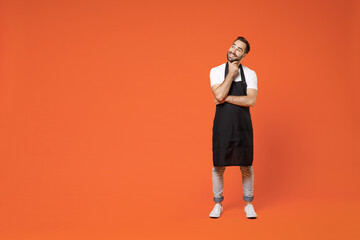 Fototapeta na wymiar Full length dreamful young man barista bartender barman employee in black apron white t-shirt work in coffee shop prop up chin look aside isolated on orange background. Small business startup concept.