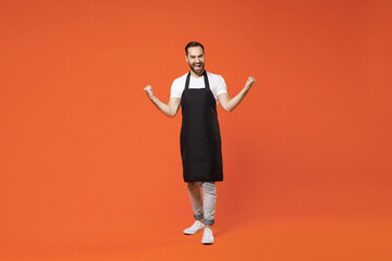 Fototapeta na wymiar Full length young fun man barista bartender barman employee in black apron white t-shirt work in coffee shop do winner gesture clench fist isolated on orange background studio Small business startup
