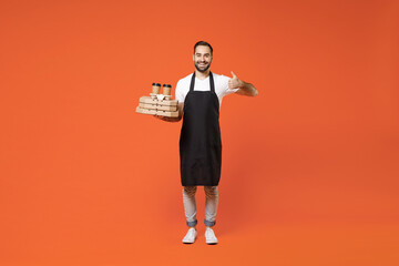 Fototapeta na wymiar Full length young man barista bartender barman employee in black apron white tshirt work coffee shop hold delivery cup pizza flatbox show thumb up isolated on orange background Small business startup