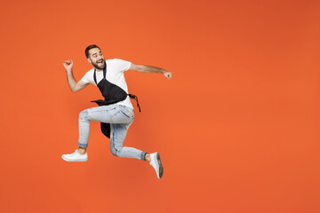 Full length side view young man barista bartender barman employee in black apron white t-shirt work in coffee shop jump high run fast isolated on orange background. Small business startup concept.