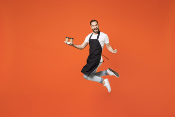 Fototapeta na wymiar Full length young man barista bartender barman employee in apron white t-shirt work in coffee shop jump hold paper delivery cup do winner gesture isolated on orange background. Small business startup.