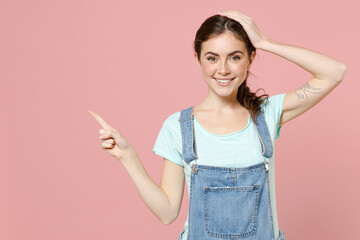 Young happy surprised caucasian woman in trendy denim clothes blue t-shirt point index finger aside on workspace area mock up copy space hold face isolated on pastel pink background studio portrait