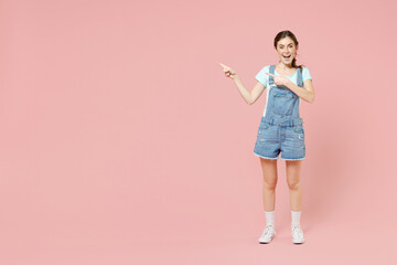 Full length young smiling happy cheerful caucasian woman 20s in trendy denim clothes blue t-shirt point index finger aside on workspace area mock up isolated on pastel pink background studio portrait