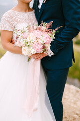 The bride and groom stand hugging and hold the bride's bouquet with delicate pink roses, peonies and astilbe, close-up 