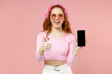 Young smiling happy woman in rose clothes bandana glasses hold mobile cell phone blank screen workspace area show thumb up gesture isolated on pastel pink background studio People lifestyle concept.