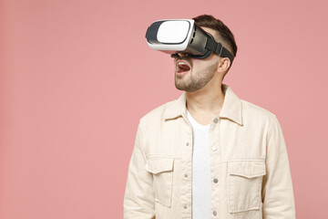 Young happy fun amazed caucasian impressed man 20s wearing jacket white t-shirt watching in vr headset pc gadget isolated on pastel pink color background studio portrait. People lifestyle concept.
