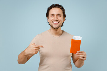 Traveler tourist man 20s in casual basic t-shirt point index finger on passport tickets isolated on pastel blue background. Passenger traveling abroad on weekends getaway. Air flight journey concept.