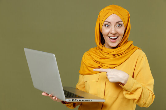 Surprised young arabian asian muslim woman in abaya hijab yellow clothes point index finget on laptop computer isolated on olive green background studio portrait. People uae islam religious concept
