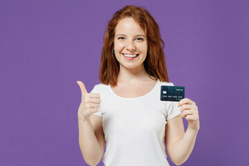 Young smiling rich successful satisfied redhead woman 20s in white basic blank print design t-shirt hold credit bank card show thumb up like gesture isolated on dark violet background studio portrait