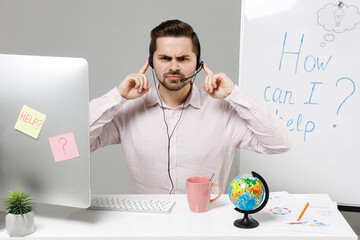 Employee angry operator business man in set microphone headset for helpline assistance sit work at call center office desk with pc computer cover ears with fingers isolated on grey background studio