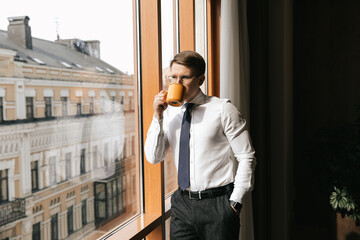Young businessman stands with a cup of coffee by the window of his office