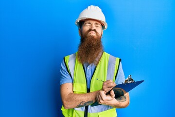 Fototapeta premium Redhead man with long beard wearing safety helmet holding clipboard looking at the camera blowing a kiss being lovely and sexy. love expression.