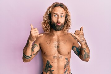 Handsome man with beard and long hair standing shirtless showing tattoos doing shaka sign looking...