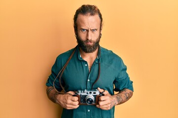 Handsome man with beard and long hair holding vintage camera skeptic and nervous, frowning upset...