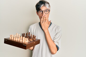 Young hispanic man holding chess covering mouth with hand, shocked and afraid for mistake....