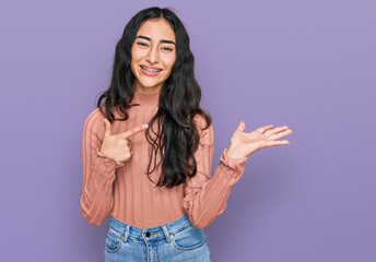 Hispanic teenager girl with dental braces wearing casual clothes amazed and smiling to the camera while presenting with hand and pointing with finger.