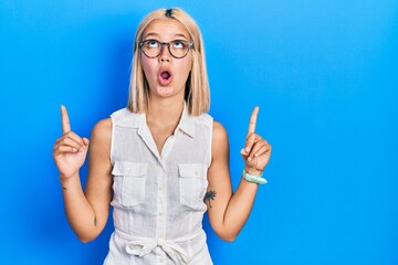 Beautiful blonde woman wearing glasses amazed and surprised looking up and pointing with fingers and raised arms.