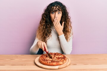 Young hispanic girl cutting tasty pepperoni pizza covering mouth with hand, shocked and afraid for...