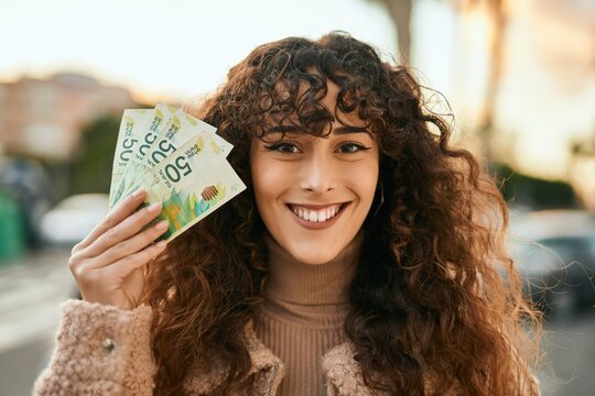 Young hispanic woman smiling happy holding israel shekels banknotes at the city