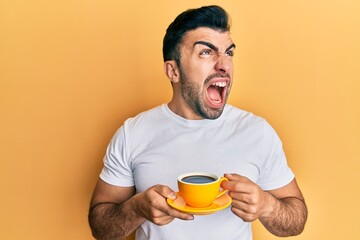 Young hispanic man drinking a cup of coffee angry and mad screaming frustrated and furious, shouting with anger looking up.