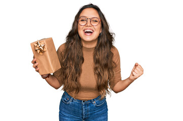 Young hispanic girl holding gift screaming proud, celebrating victory and success very excited with...