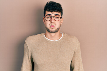 Young hispanic man wearing casual clothes and glasses puffing cheeks with funny face. mouth...