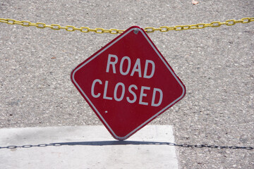 Diamond shaped red ROAD CLOSED sign on a yellow chain over a city street close to the ground blocking off traffic