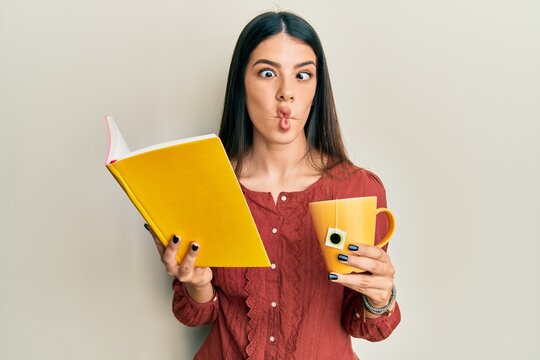 Young hispanic woman reading book drinking cup of tea making fish face with mouth and squinting eyes, crazy and comical.