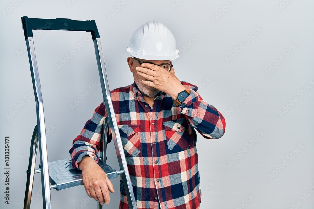 Sticker Handsome mature handyman close to construction stairs wearing hardhat covering eyes with hand, looking serious and sad. sightless, hiding and rejection concept