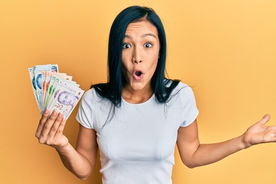 Beautiful hispanic woman holding singapore dollars banknotes celebrating achievement with happy smile and winner expression with raised hand