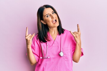 Middle age brunette woman wearing doctor uniform and stethoscope shouting with crazy expression doing rock symbol with hands up. music star. heavy concept.