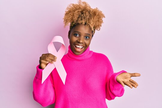 Young african woman with afro hair holding pink cancer ribbon celebrating achievement with happy smile and winner expression with raised hand