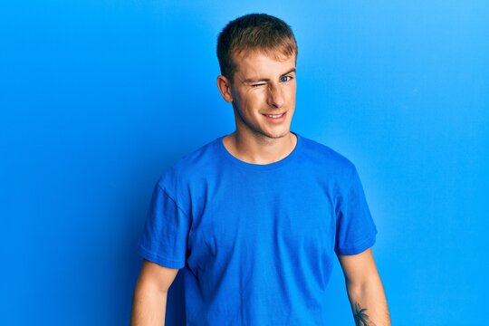 Young caucasian man wearing casual blue t shirt winking looking at the camera with sexy expression, cheerful and happy face.