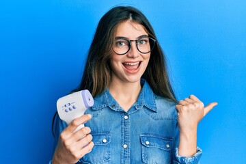 Young caucasian woman holding digital thermometer pointing thumb up to the side smiling happy with...