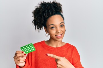 Young african american girl holding birth control pills smiling happy pointing with hand and finger