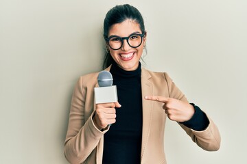 Young hispanic woman holding reporter microphone smiling happy pointing with hand and finger