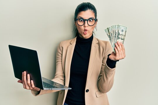 Young hispanic woman wearing business style holding laptop and 50 euros making fish face with mouth and squinting eyes, crazy and comical.