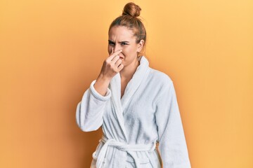 Young blonde woman wearing bathrobe smelling something stinky and disgusting, intolerable smell,...
