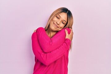 Fototapeta premium Beautiful hispanic woman wearing casual pink sweater hugging oneself happy and positive, smiling confident. self love and self care