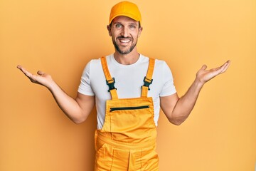 Young handsome man wearing handyman uniform over yellow background smiling showing both hands open...