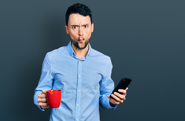 Hispanic man with beard using smartphone and drinking a cup of coffee making fish face with mouth and squinting eyes, crazy and comical.