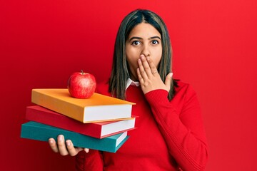 Young latin woman holding books and apple covering mouth with hand, shocked and afraid for mistake. surprised expression