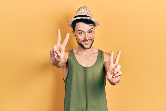 Young hispanic man wearing summer hat smiling looking to the camera showing fingers doing victory sign. number two.