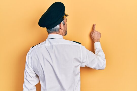 Handsome middle age man with grey hair wearing airplane pilot uniform posing backwards pointing ahead with finger hand