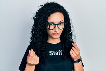 Young hispanic woman with curly hair wearing staff t shirt doing money gesture with hands, asking for salary payment, millionaire business